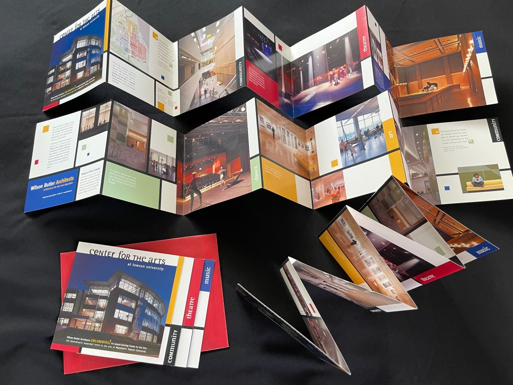 Photo of a colorful folding brochure with images and text describing the opening of the Center for the Arts building. The brochure is shown lying flat on a red mailing envelope, standing up, and open/extended.